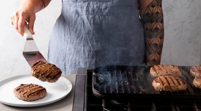 Hamburger patty is transferred to a plate with another patty using a metal offset turner. Four other patties continue to cook on a stovetop griddle. 